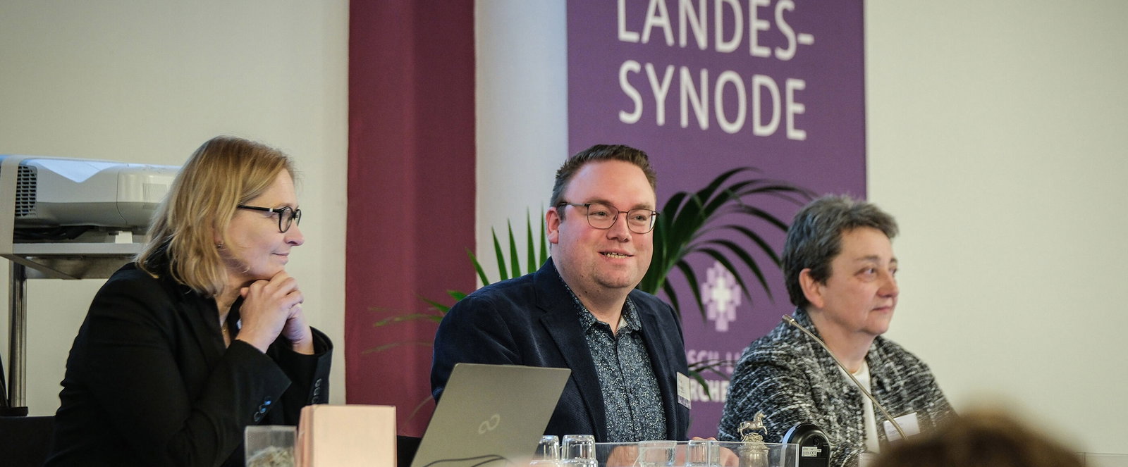 Eine männlich und zwei weiblich zu lesende Personen sitzen an einem Pult.