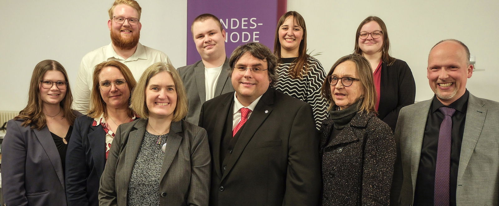 Sechs weiblich und vier männlich zu lesende Personen stehen als Gruppe zusammen und schauen in die Kamera.