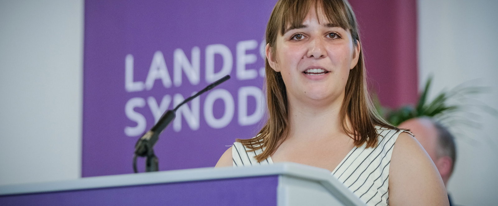 Eine Frau in einem gestreiften Kleid steht hinter einem Rednerpult, hinter ihr ein lilafarbener Banner auf dem Landessynode steht.