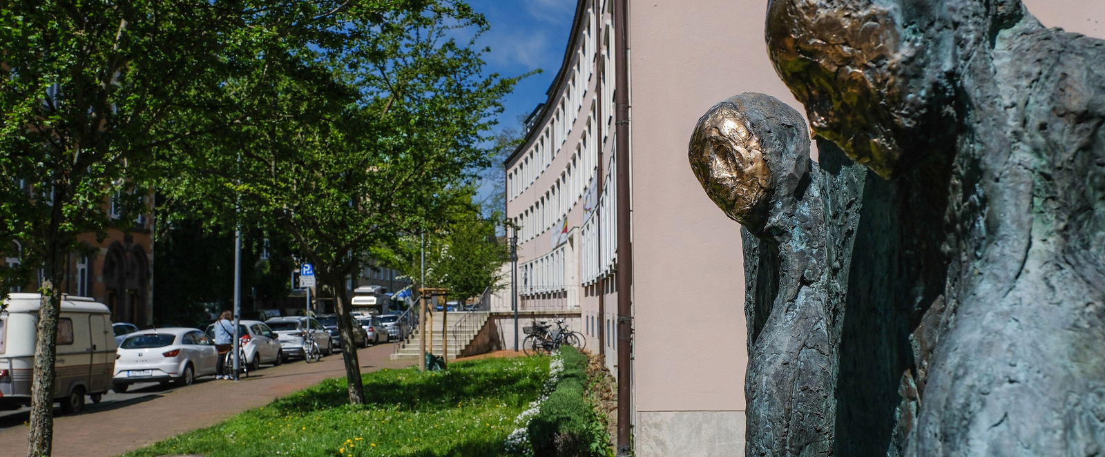 Das Bild zeigt zwei grau-goldene Skulpturen im Vordergrund vor einem rosafarbenen Gebäude an einer Straße mit belaubten Bäumen.