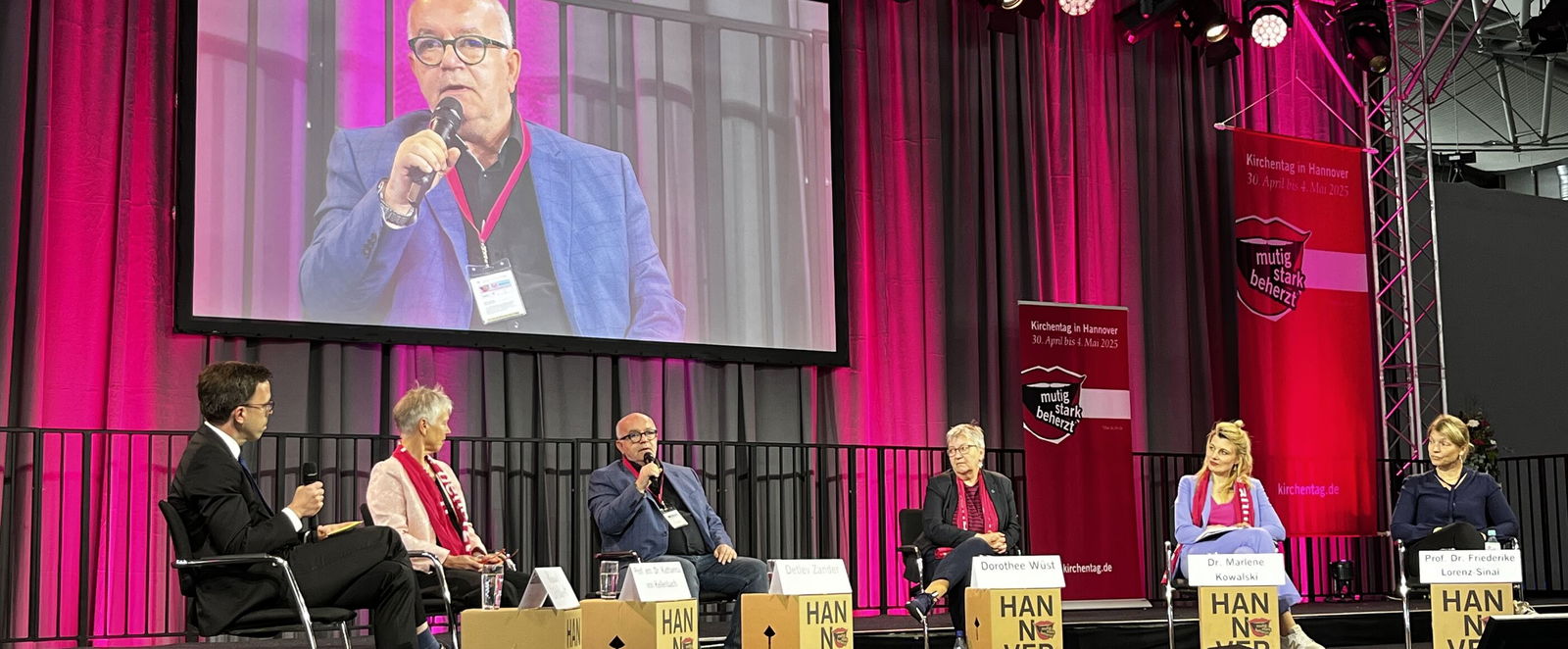 auf einem Podium sitzen sechs Personen im Halbkreis; im Hintergrund hängt ein Bildschirm mit einer männlich gelesenen Person am Mikro