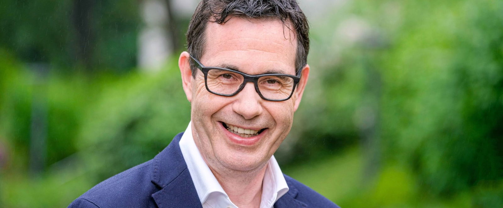 Auf dem Bild ist ein Portrait von Stephan Schaede zu sehen. Er trägt ein blaues Jackett und ein weisses Hemd. Stephan Schaede hat kurze, braune Haare, und er traegt eine Brille.