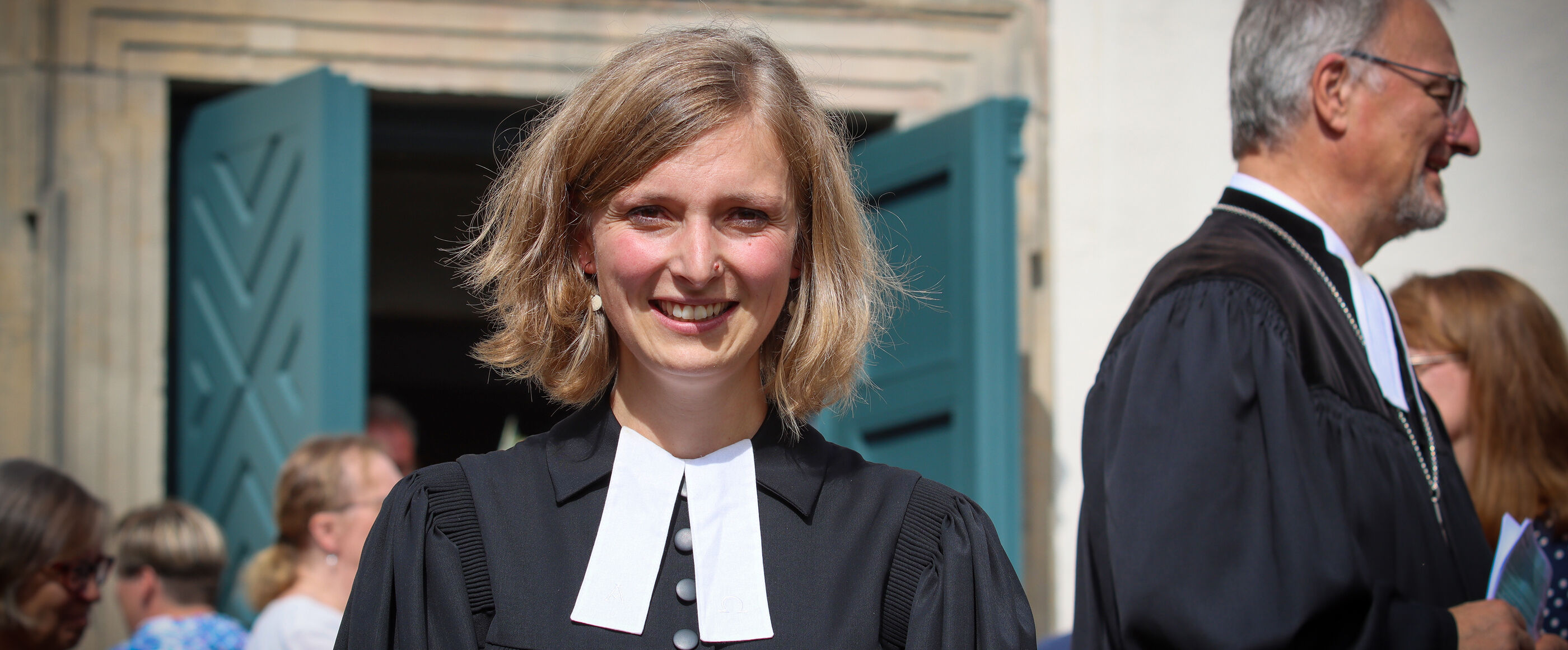 eine junge Pastorin steht vor einer Kirche. Sie hat blonde Haare, lächelt in die Kamera und trägt einen schwarzen Talar.