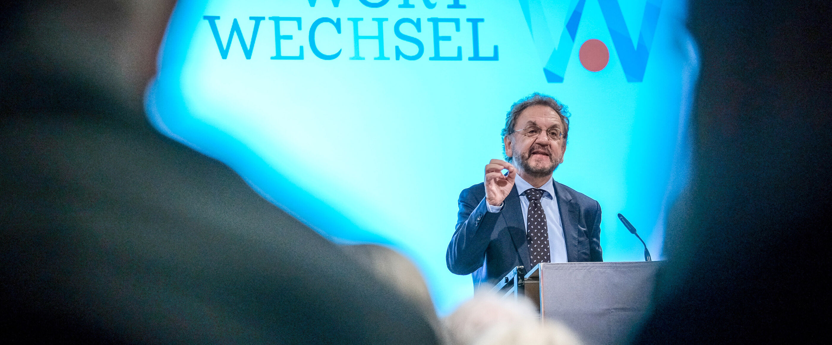 Ein Mann im Anzug spricht an einem Redepult, im Hintergrund auf der Leinwand steht „Wortwechsel“.
