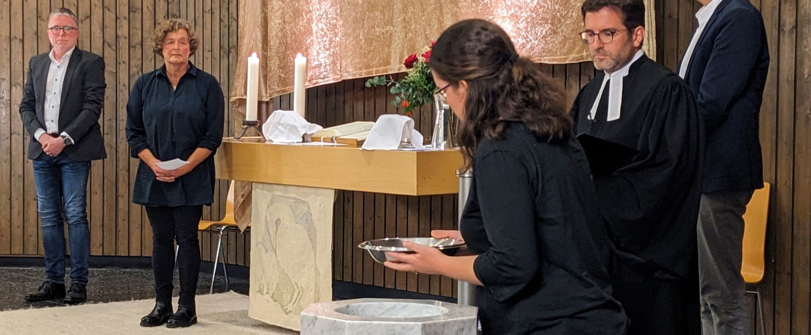 Eine Frau nimmt eine Taufschale aus einem Taufstein, vier weitere Personen stehen dabei.