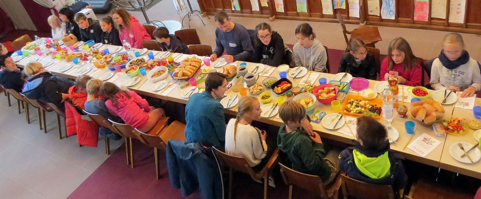 Viele Kinder sitzen mit einigen Erwachsenen an einem langen gedeckten Tisch in einer Kirche.