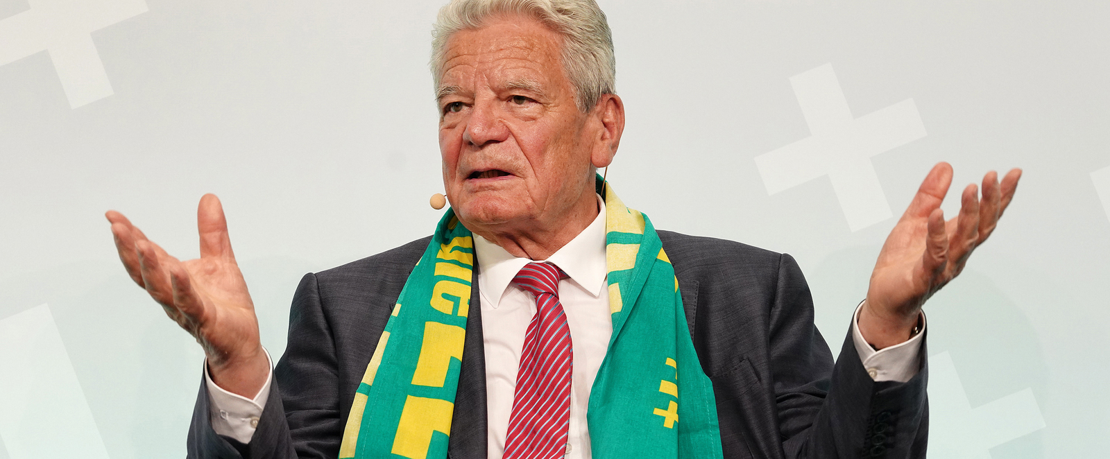 Ein älterer Mann mit grauem Haar und Kirchentagsschal spricht und hebt dabei beide Hände.