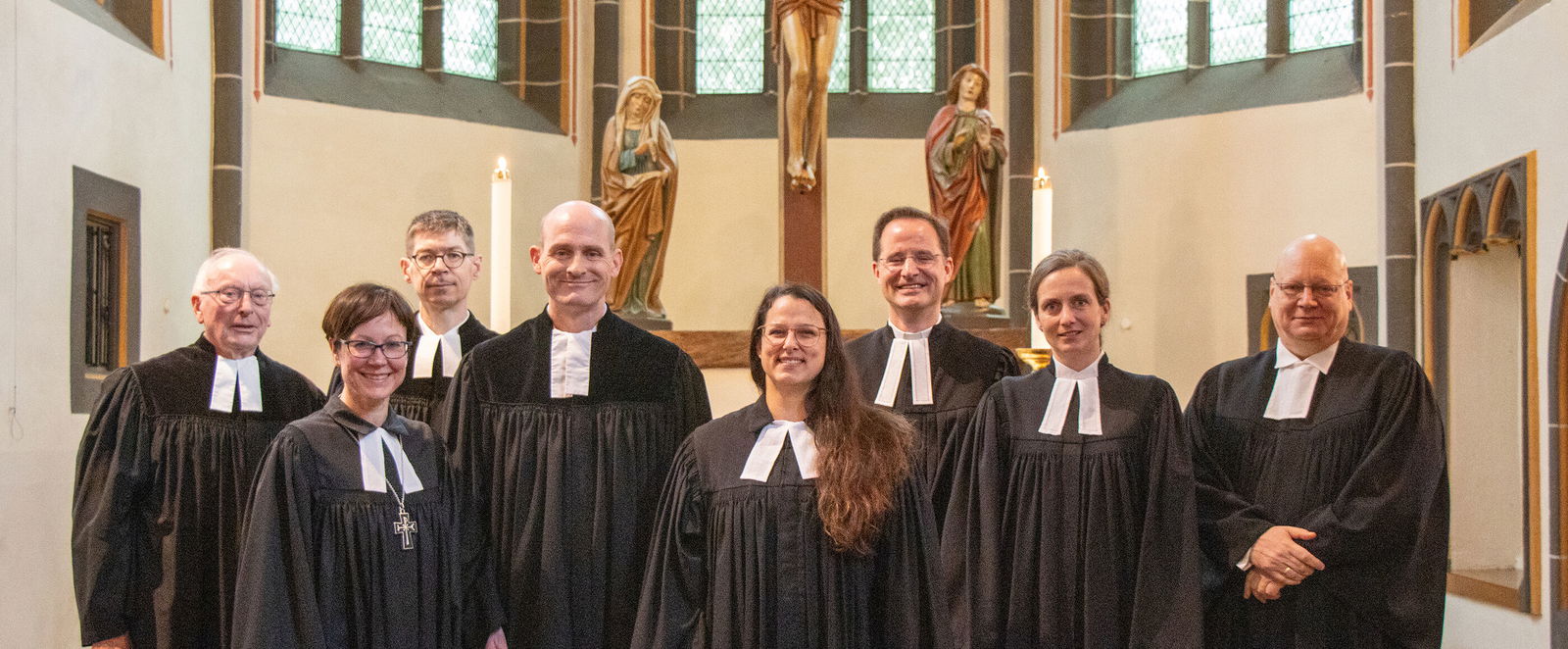 Acht Personen in Talaren stehen zusammen im Altarraum einer Kirche, über ihnen hängt ein großes Kruzifix. 
