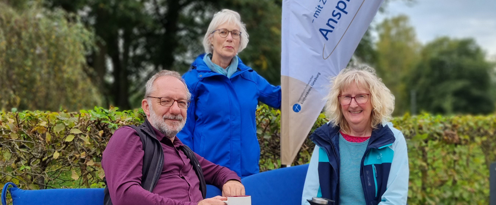 Ein Mann und eine Frau sitzen auf einer Bank mit blauen Sitzkissen. Hinter ihnen steht eine Frau in blauer Jacke. Im Hintergrund eine Hecke.