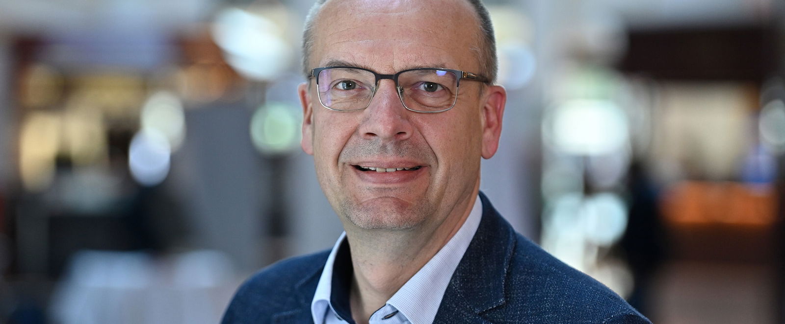 Eine als Mann lesbare Person im hellblauen Hemd und mit Sakko lächelt in die Kamera.