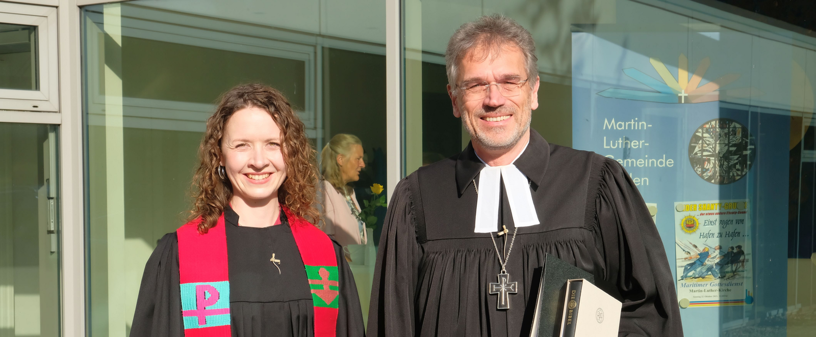 Eine junge Frau steht neben einem älteren Mann, beide tragen schwarze Talare. Die Frau trägt darüber eine bunte Stola. 