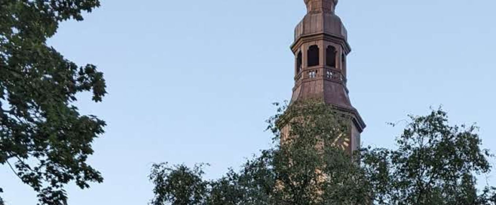 Über grüne Baumwipfel ragt ein Kirchturm in den blauen Himmel.