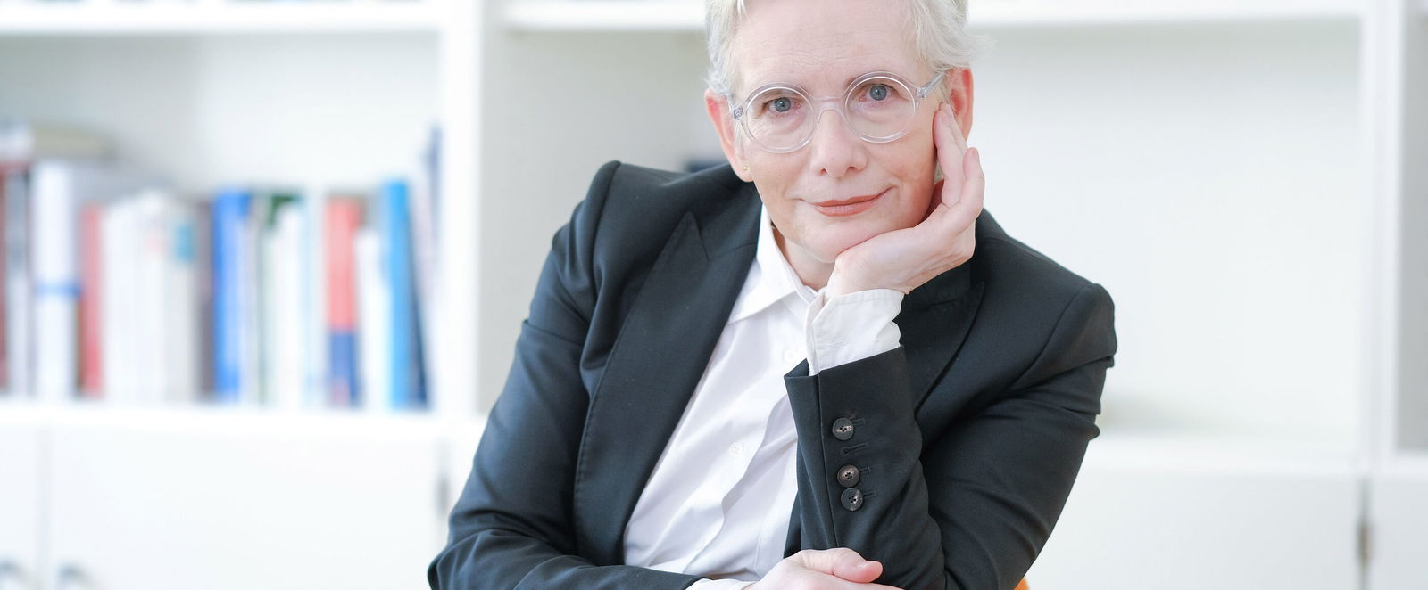 Eine Frau mit sehr hellen Haaren und Brille und dunklem Jackett sitzt in einem orangenen Sessel vor Bücherregalen.