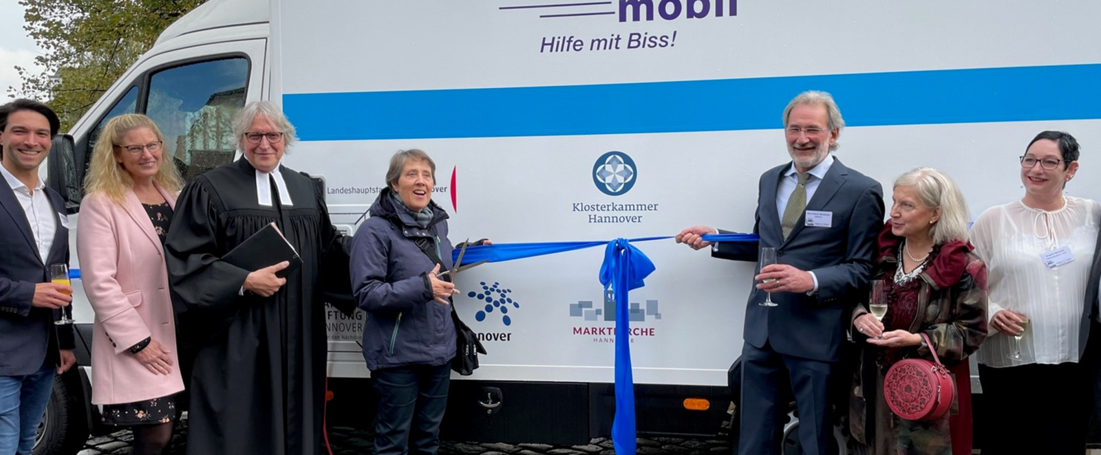Vor einem weiß-blauen Kastenwagen, auf dem "Zahnmobil" steht, stehen mehrere lachende Menschen. Zwei halten eine blaue Schleife.