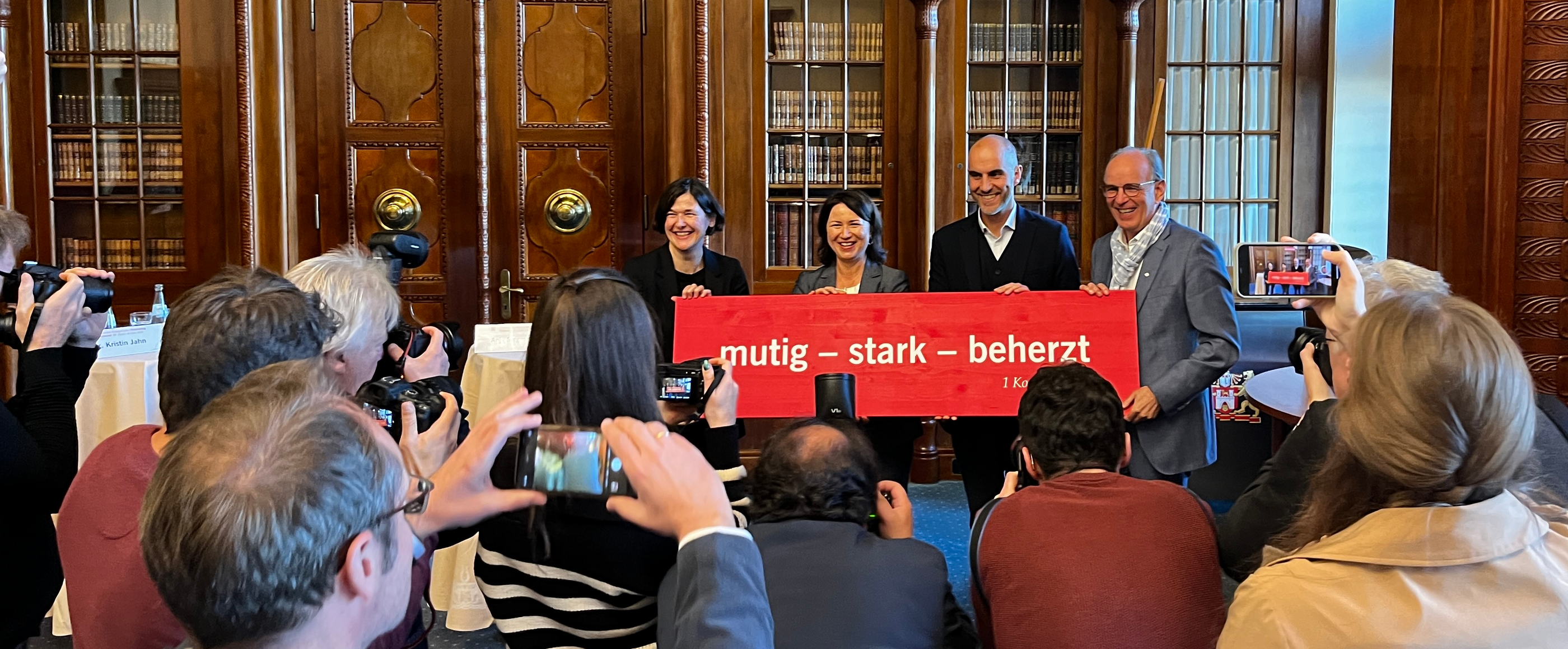 Vier Menschen halten ein rotes Banner auf dem steht weiß: Mutig. stark. beherzt. Vor ihnen stehen andere Menschen ihnen gegenüber, manche halten Smartphone in die Luft wie zum Fotografieren.