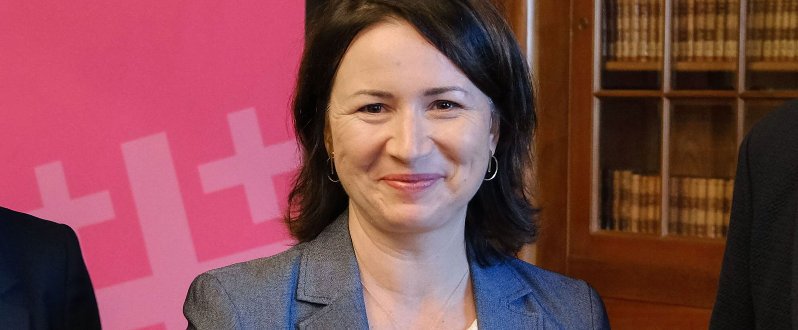 Eine Frau mit mittellangen, dunklen Haaren und grauem Blazer steht vor einem dunklen Schrank mit Glaseinsätzen, durch die Bücher erkennbar sind. Links steht ein rosa Aufsteller mit der weißen Aufschrift "Kirchentag 30. April bis 4. Mai 2025 in Hannover"