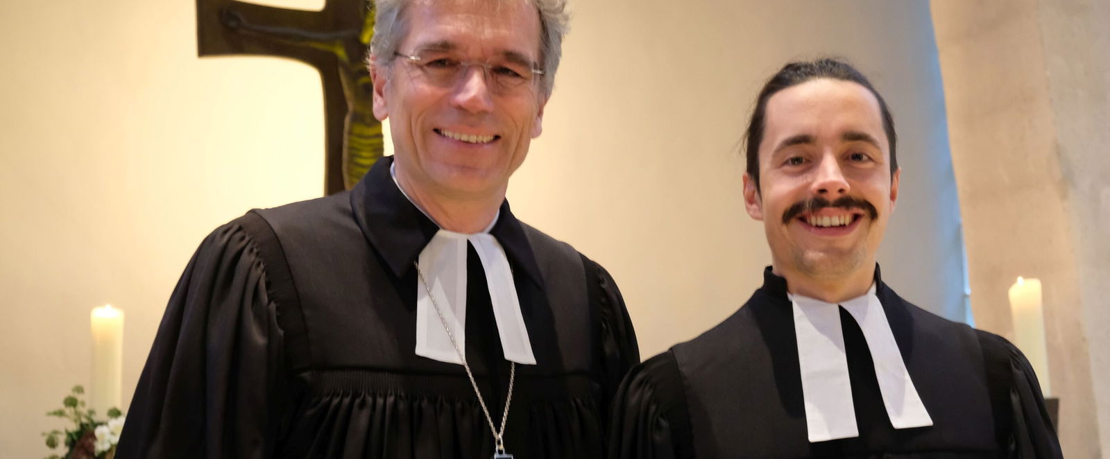 Zwei Pastoren in schwarzen Talaren stehen vor einem Kreuz. Links und rechts sind weiße Altarkerzen zu sehen