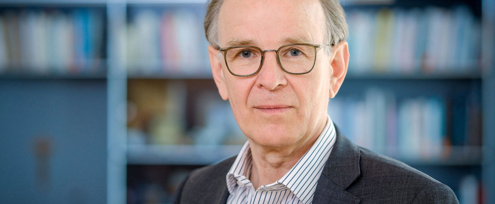 Ein Mann mit schütterem Haar, Brille, Sakko, gestreiftem Hemd und ernstem Blick vor einer Bücherwand.