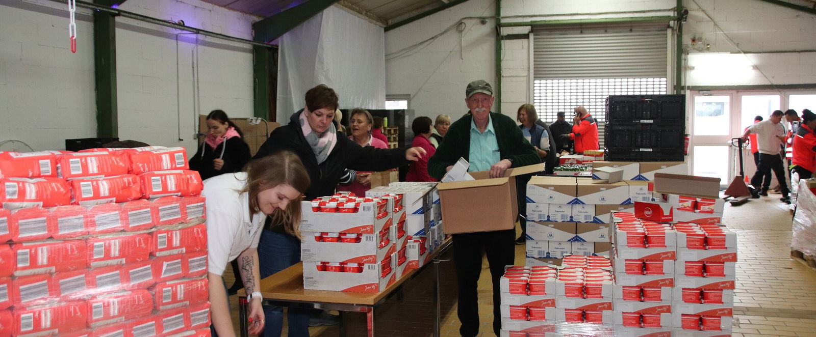 In einer Lagerhalle stehen Paletten voll Mehl und Zucker, einige Personen packen daraus kleinere Pakete an langen Tischen.