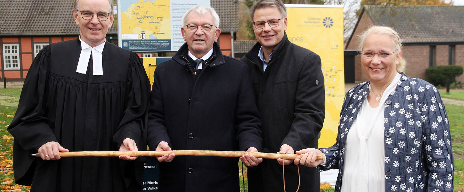 Drei Männer und eine Frau halten einen Wanderstab vor einer Infosäule.