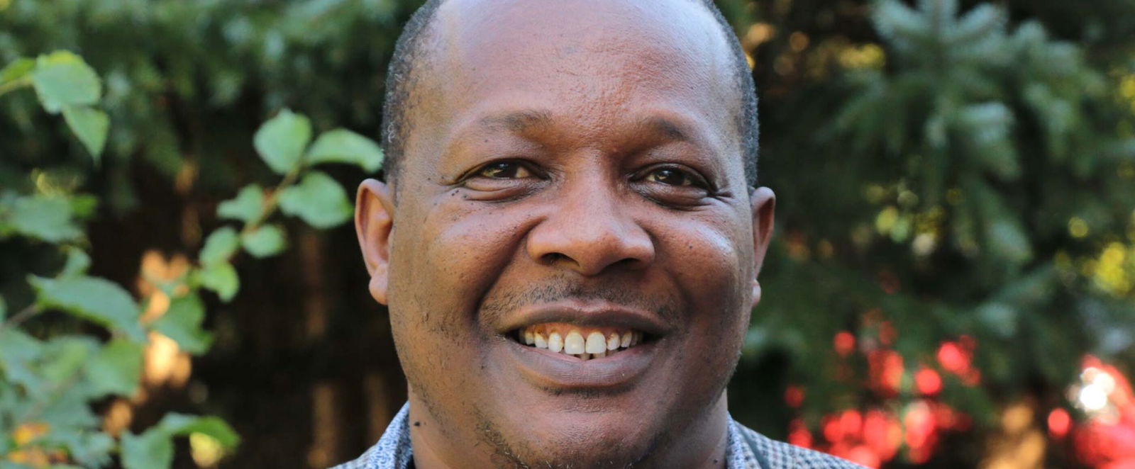 Ein schwarzer Mann steht lächelnd im Grünen. Er trägt ein graues Sakko und ein hellblaues Hemd.