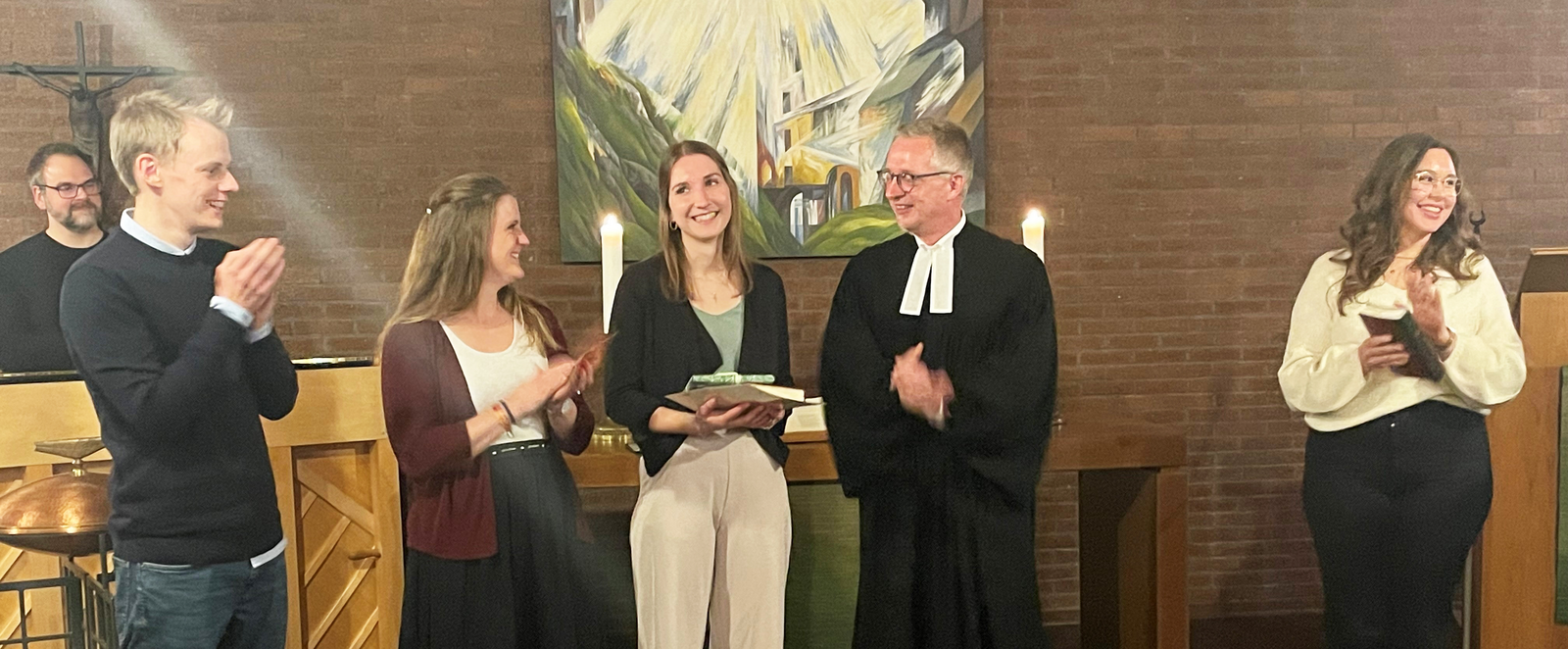 Mehrere Personen stehen nebeneinander, alle bis auf eine klatschen. Diese hält eine Mappe in Händen. Eine Person trägt einen Talar.