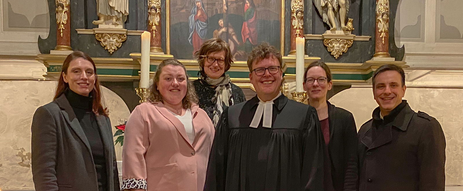 Sechs Personen stehen vor einem Altar, eine trägt einen Talar.