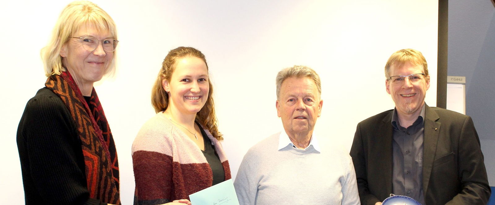 Zwei Frauen und zwei Männer stehen nebeneinander. Eine Frau hält einen Zettel in der Hand, alle lächeln.