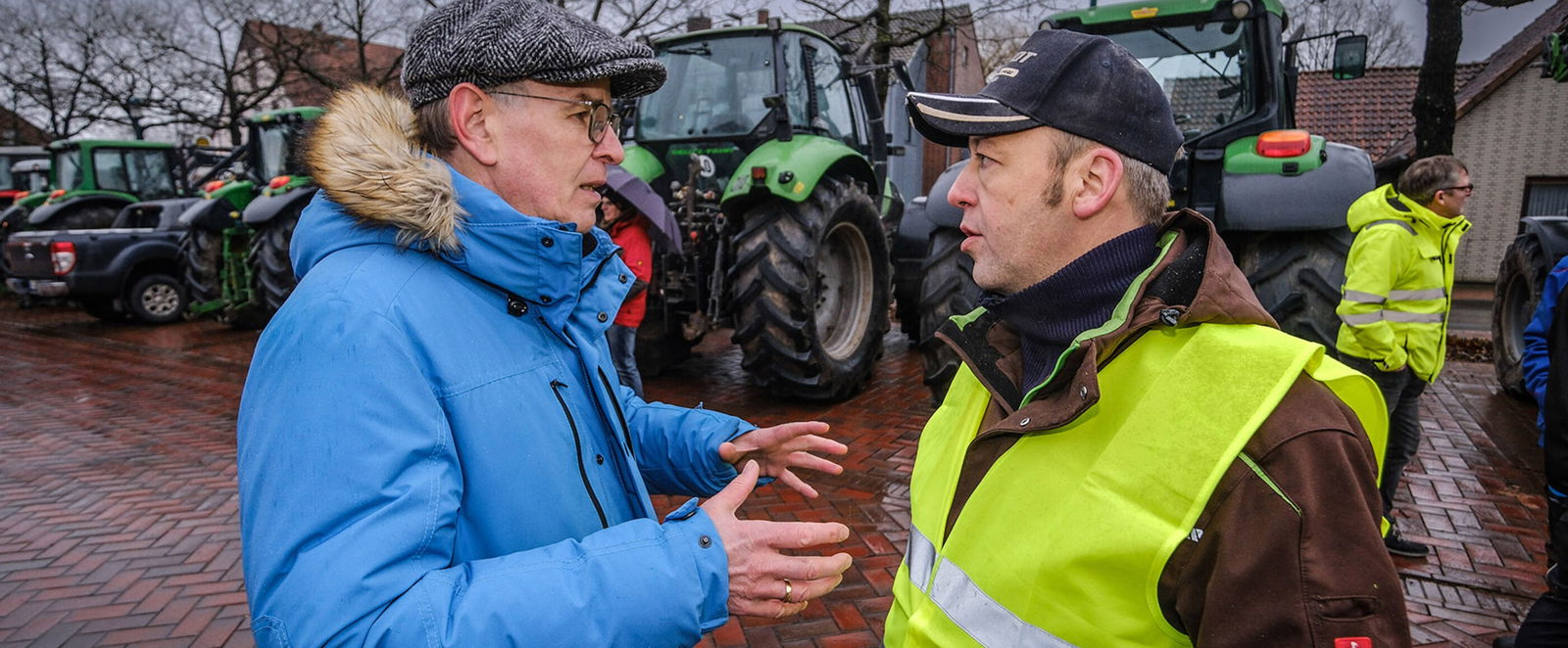 Zwei Männer, einer mit blauer, einer mit gelber Warnweste über einer braunen Jacke, beide mit Mützen, stehen sich gegenüber, der links gestikuliert. Im Hintergrund stehen Traktoren.
