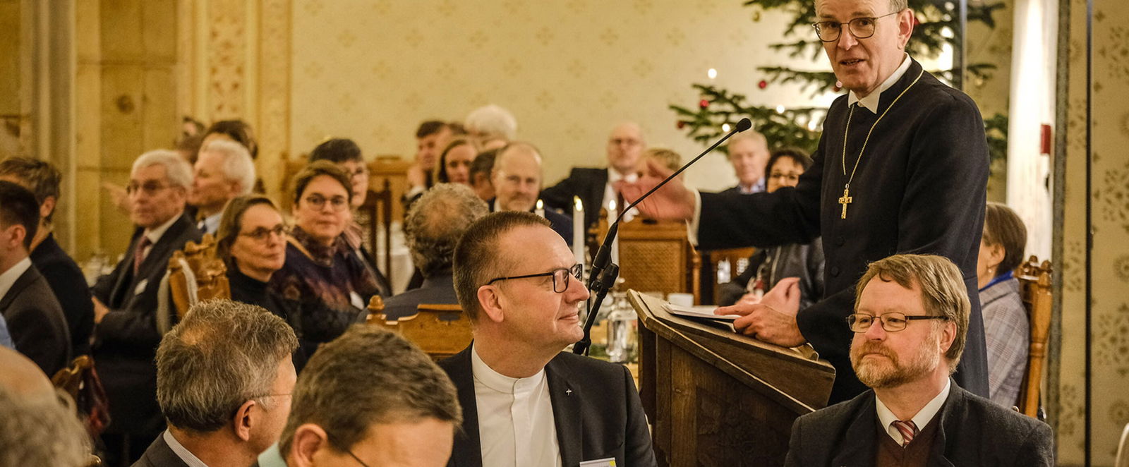 Viele Menschen sitzen an mehreren Tischen zusammen, einer steht zwischen ihnen, mit schwarzer Robe und Kreuz um den Hals. Im Hintergrund ein Weihnachtsbaum.