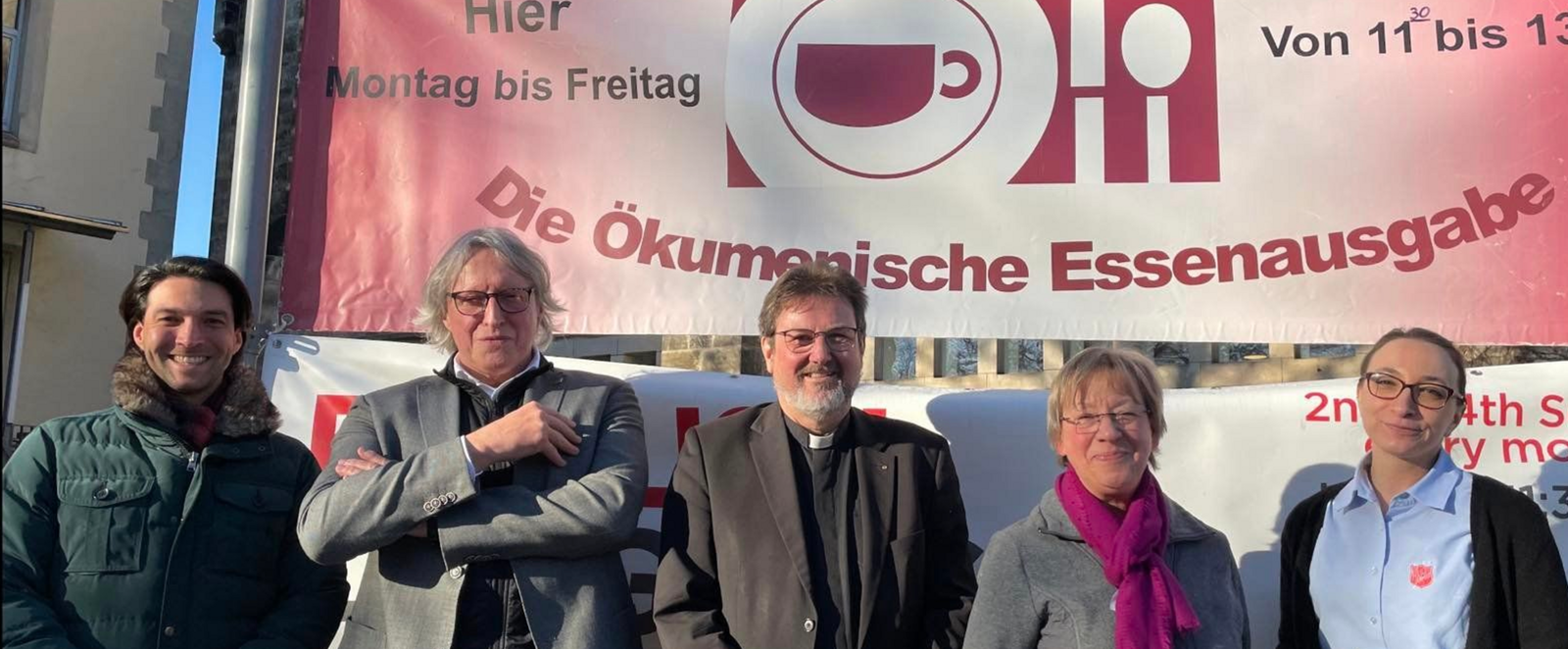Fünf Menschen stehen in Reihe vor einem roten Plakat, auf dem steht: Ökumenische Essensausgabe, Montag bis Freitag, 11 bis 13 Uhr. 