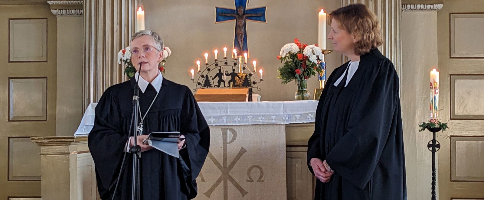 Zwei Frauen in Talaren stehen vor einem Altar mit Kanzel darüber.