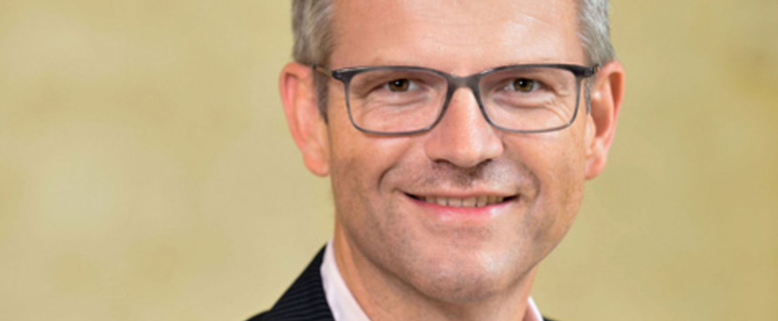 Ein Mann mit kurzen grauen Haaren und Brille, rosa Hemd und dunklem Jackett darüber, lächelt.