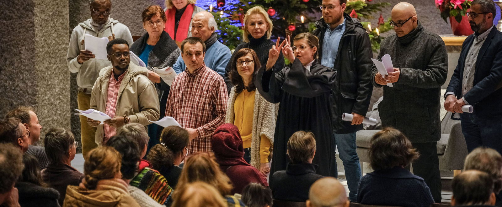Mehrere Menschen, offenbar unterschiedlicher Herkunft, stehen nebeneinander auf den Stufen vor einem Altarraum. Vor ihnen sitzen dutzende Menschen.