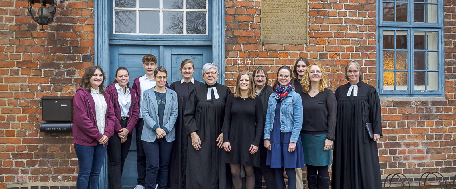 Gruppe von Menschen vor einem Backsteingebäude mit grüner Türe. 12 Personen, zwei davon tragen einen Talar.