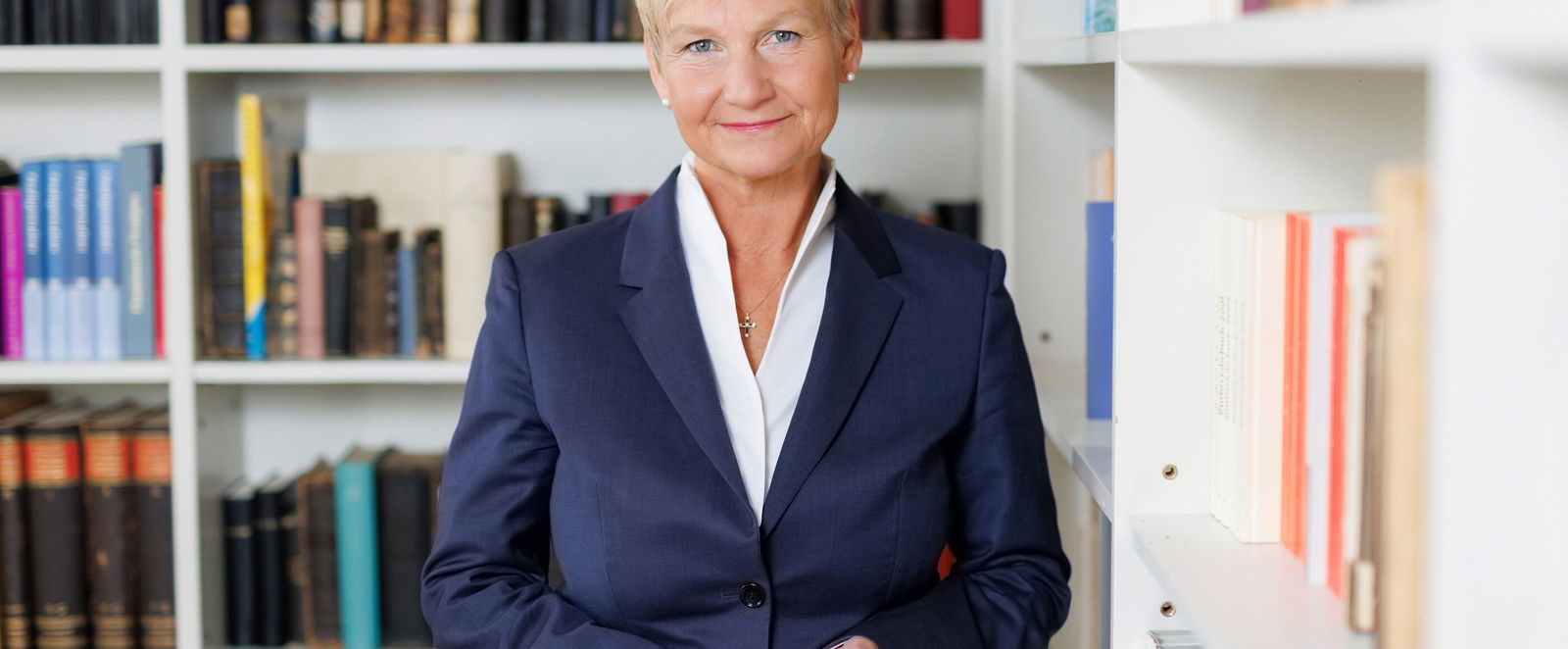 Eine als Frau lesbare Person steht vor einer Bücherwand.