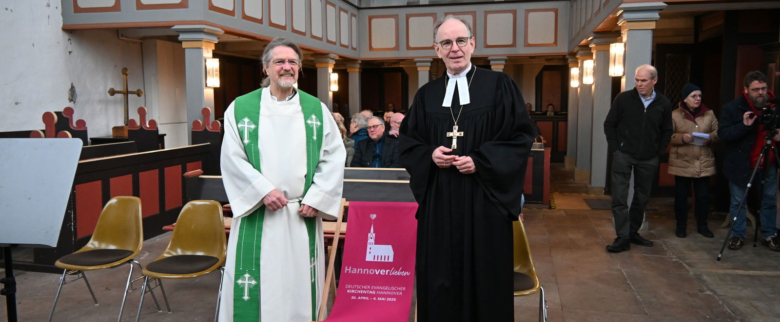 Zwei Männer,, einer im weißen, einer im schwarzen Talar, stehen links und recht eines rot-rosa Liegestuhls. Im Hintergrund eine Kirche, in deren Bänke Menschen sitzen.