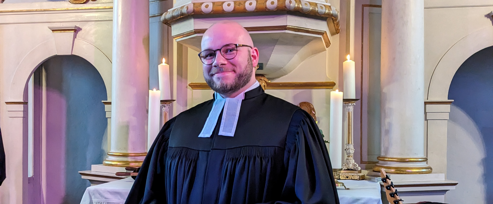Ein Mann in Talar steht in einer hellen Kirche vor dem Altar mit rotem Parament. Er hält ein Buch in den Händen.