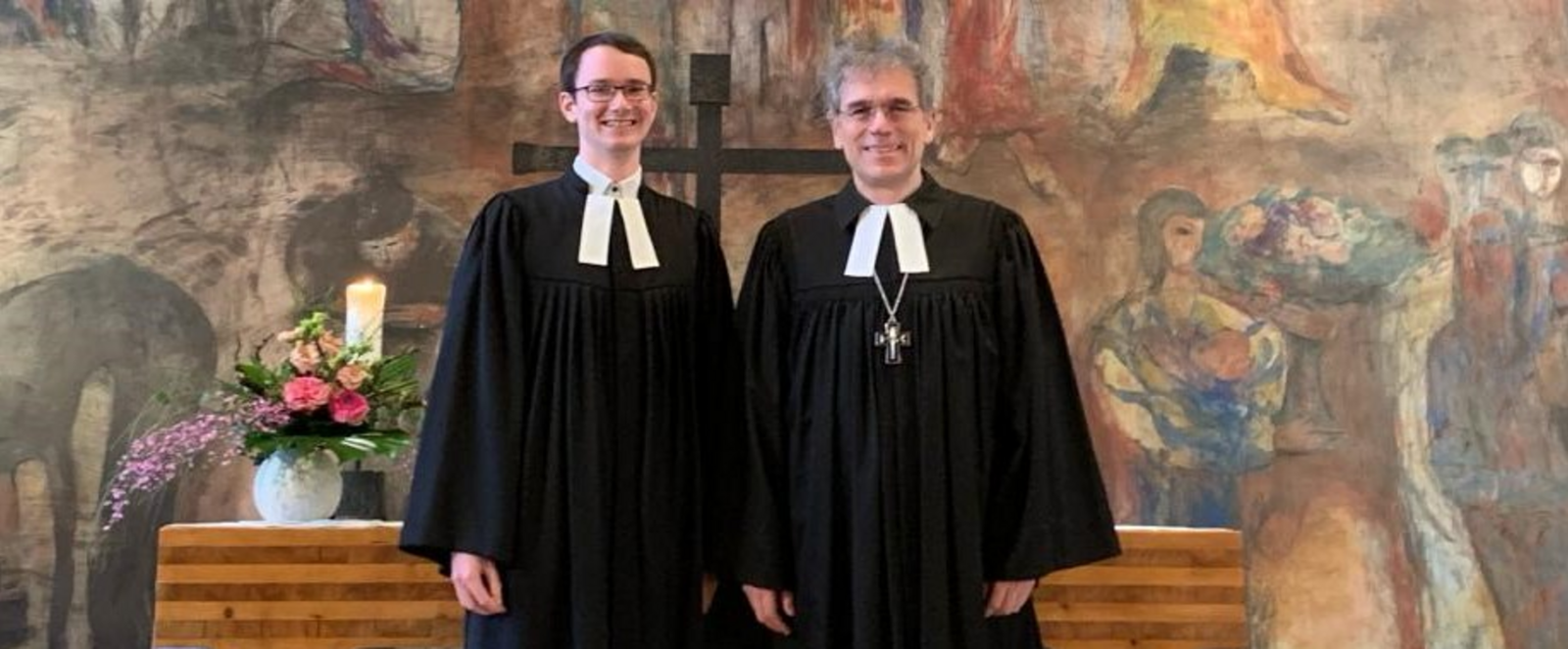 Zwei Männer in Talar stehen vor einem hölzernen Altar und lächeln in die Kamera. Im Hintergrund ist ein buntes Wandgemälde zu sehen.