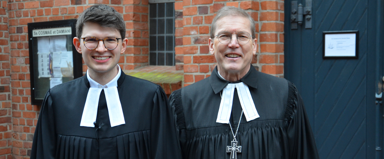 Zwei Männer tragen Talar, lächeln in die Kamera. Sie stehen vor einem Backsteingebäude mit dunkler Holztüre.