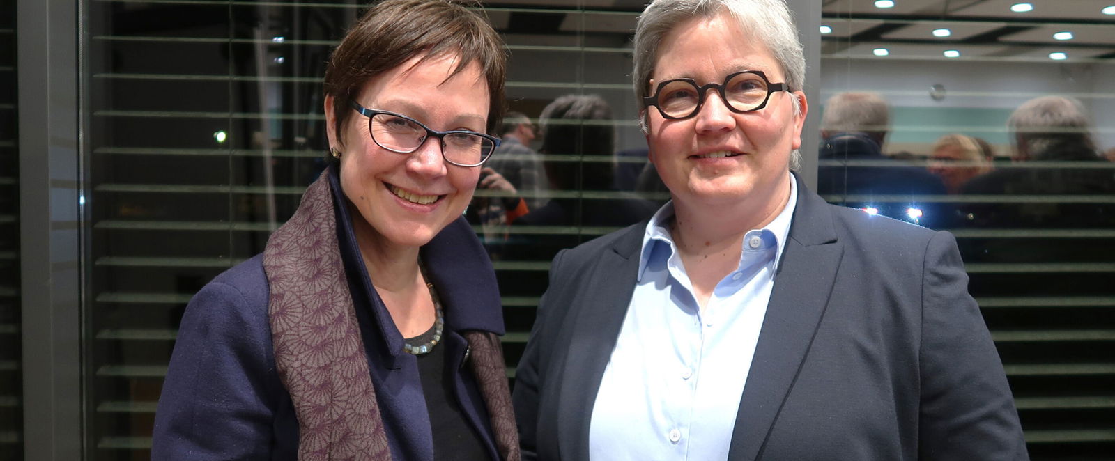 Zwei als Frauen lesbare Personen stehen vor einer Glastür und schauen in die Kamera.