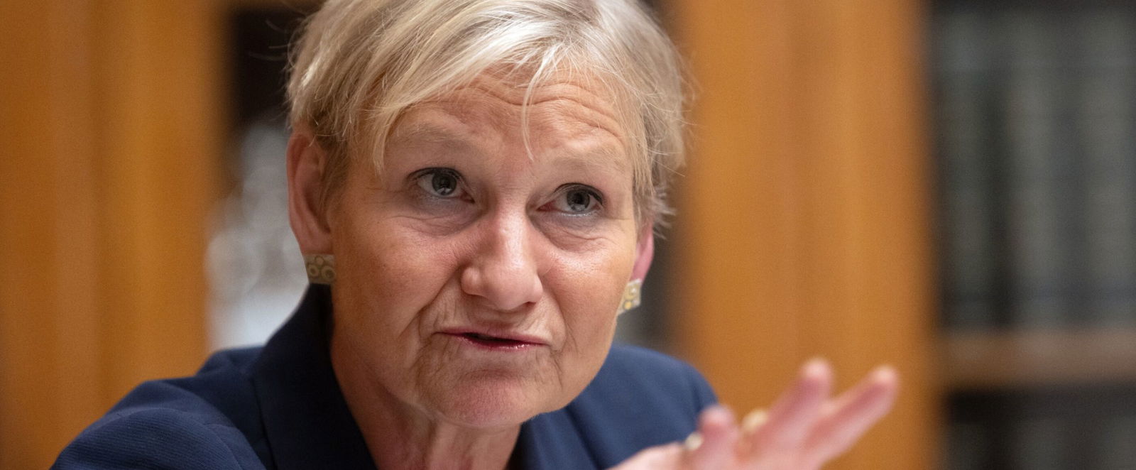 Eine Frau schaut mahnend nach vorne. Sie sitzt vor einem braunen Schrank