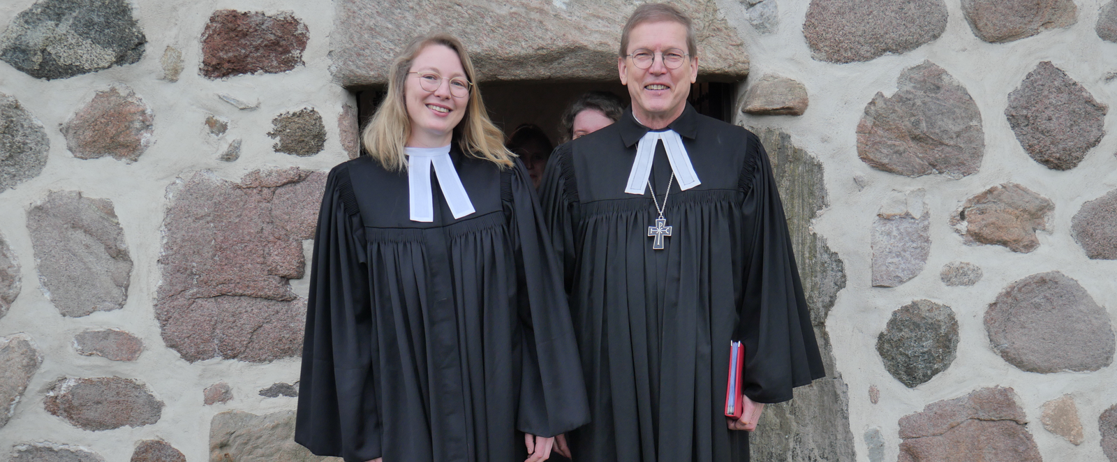 Vor einer gemauerten Fassade treten aus einer Türe eine junge frau mit blonden Haaren und Brille und neben ihr ein Mann mit kurzen braunen Haaren und Brille. Beide tragen Talar.