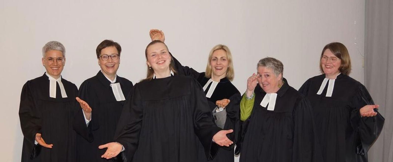 Sechs Personen in Talaren stehen zusammen und machen Gesten mit den Handflächen nach oben, nach dem Motto: "Hier sind wir"