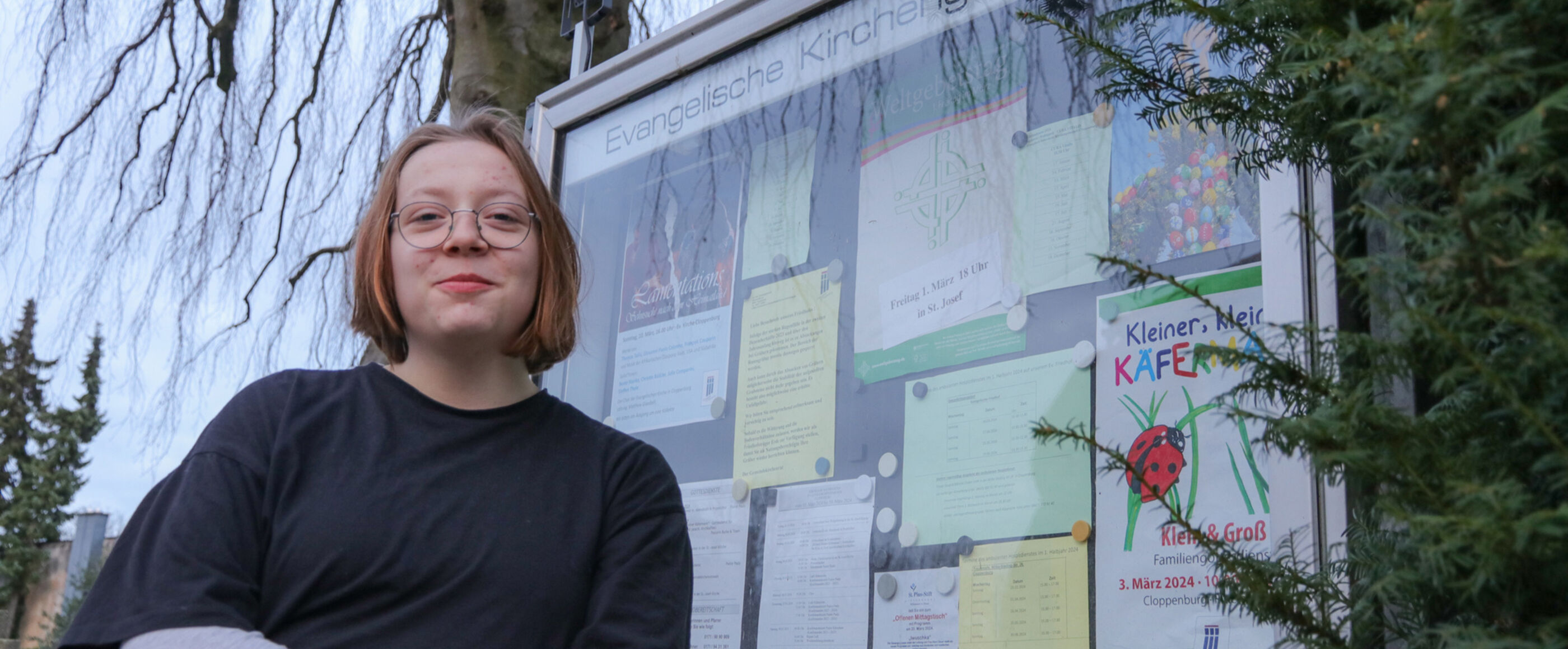 Eine junge als Frau lesbare Person steht vor dem Schaukasten ihrer Kirchengemeinde.