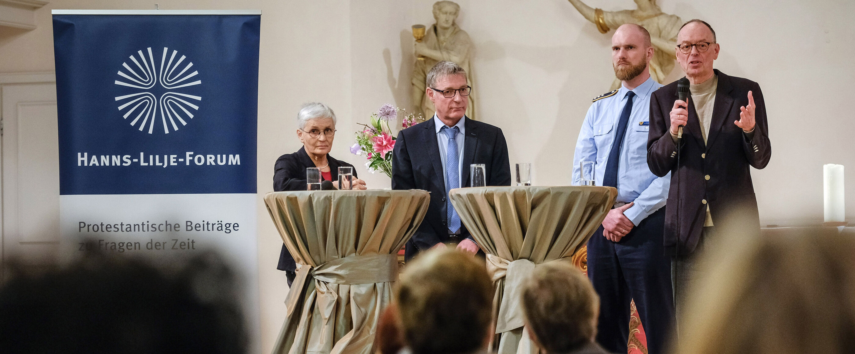 Eine Frau und drei Männer (einer davon in Uniform) stehen an Stehtischen und diskutieren.