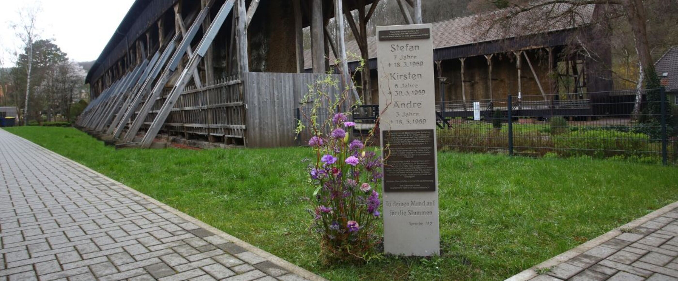 Eine steinerne Stele mit Namen drauf steht auf einer grünen Wiese. Im Hintergrund sind Gebäude aus Holz zu erkennen.