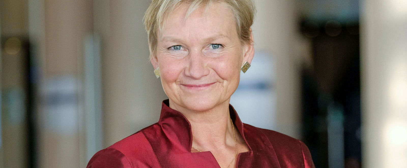 Eine weiblich gelesene Person mit kurzen, blonden Haaren und in satin-rotem Oberteil lächelt fröhlich. 