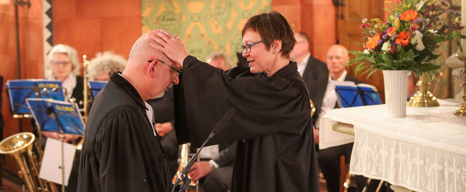 Eine weiblich gelesene Person legt einer männlich gelesenen Person die Hände auf die quasi-Glatze. Beide Tragen Talar. Im Hintergrund sitzen Menschen an Notenständern im Altarraum einer Kirche.