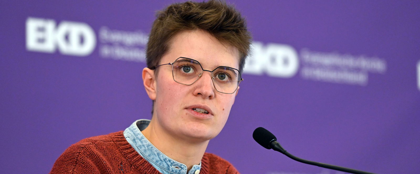 Eine junge als Frau lesbare Person mit Kurzhaarschnitt, Brille und rotem Strickpullover spricht in ein Mikrophon.